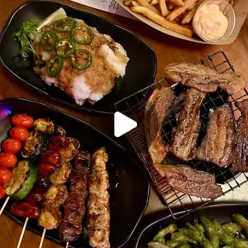 A table spread: fries with dipping sauce, a fried dish topped with sliced jalapeno, grilled skewers, sliced beef on a rack and a bowl of edamame.